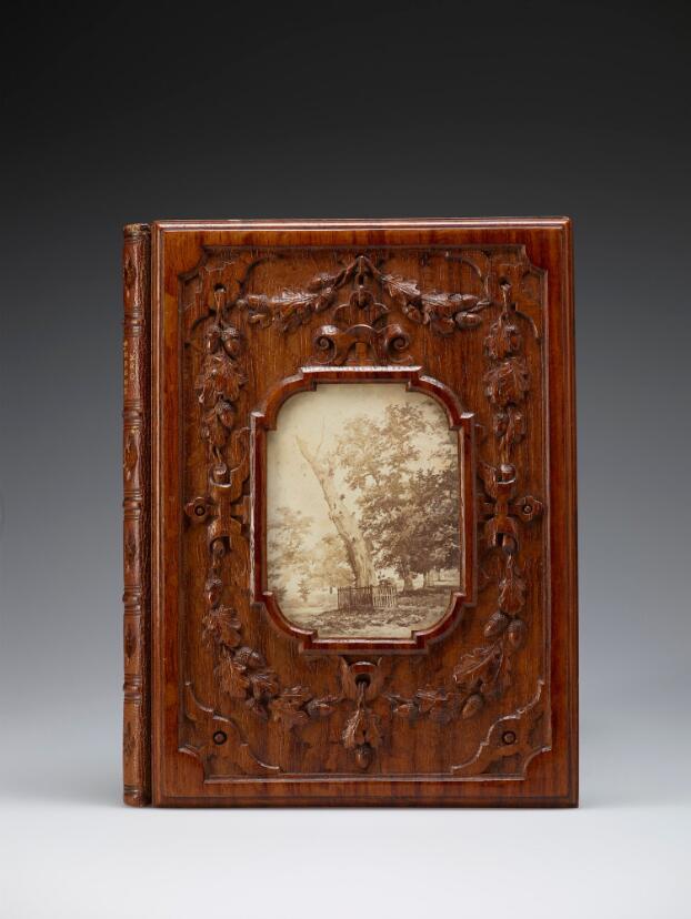 The front cover of a book with a wooden binding. The wood is varnished to a dark reddish brown, and carved with oak leaves and acorns. The title 'Herne's Oak' is just visible embossed on the spine. In the centre, a cutaway section in the wooden cover reveals a sepia photograph of an old, dying oak tree with a fence around its base, with other, living trees in the background.